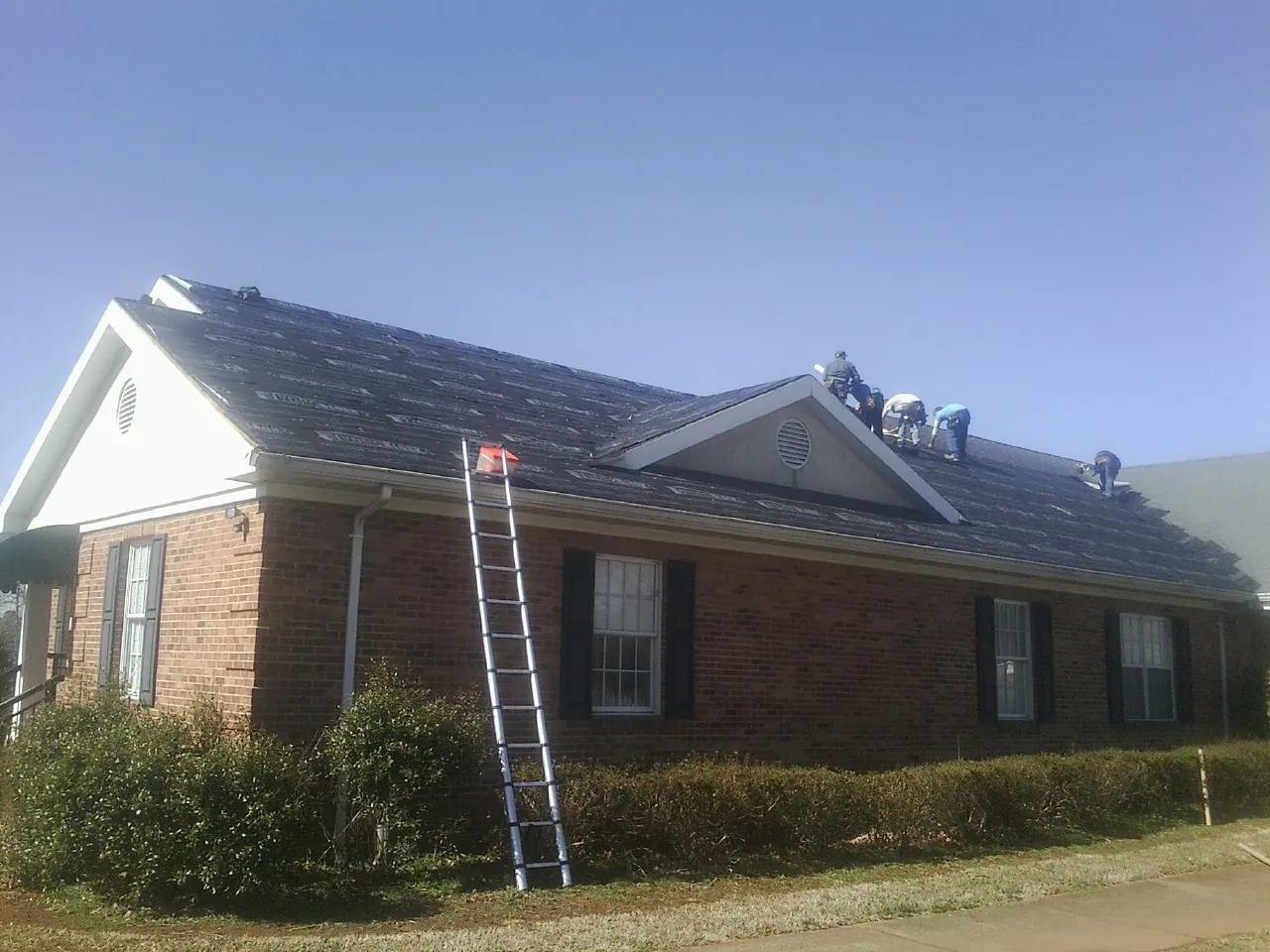 Crew tearing shingles off a brick ranch roof with a ladder set — Emergency Roof Repair work by our crew in Gurley