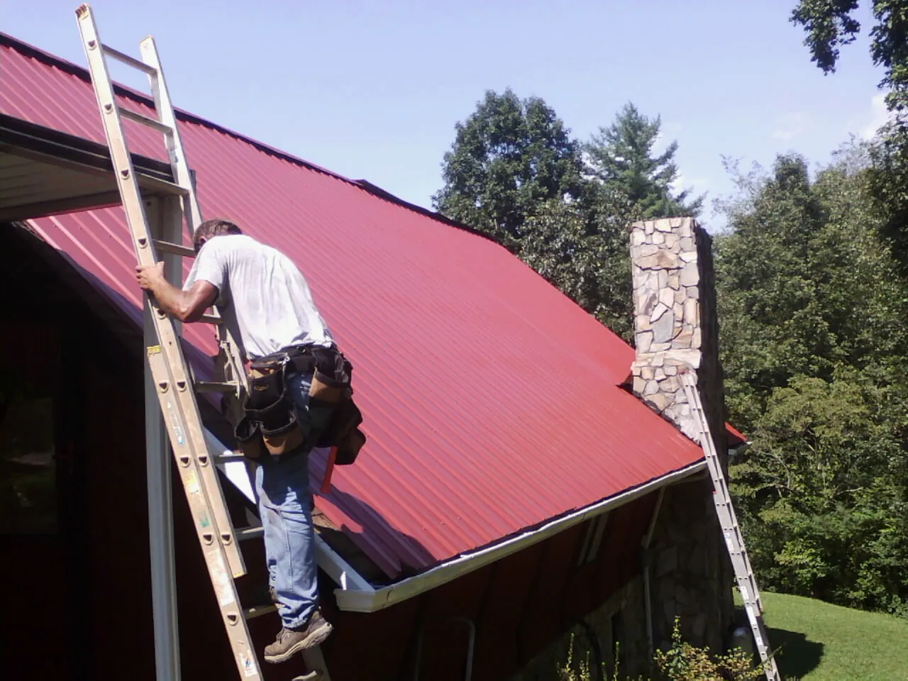 Roofer on a ladder beside a red metal roof and stone chimney — Soffit & Fascia Repair by Roofing Brothers in Gurley